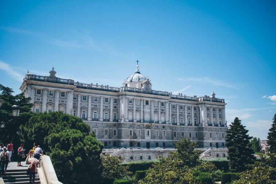 Vista frontal del Palacio Real de Madrid en día soleado, con jardines y visitantes en primer plano.